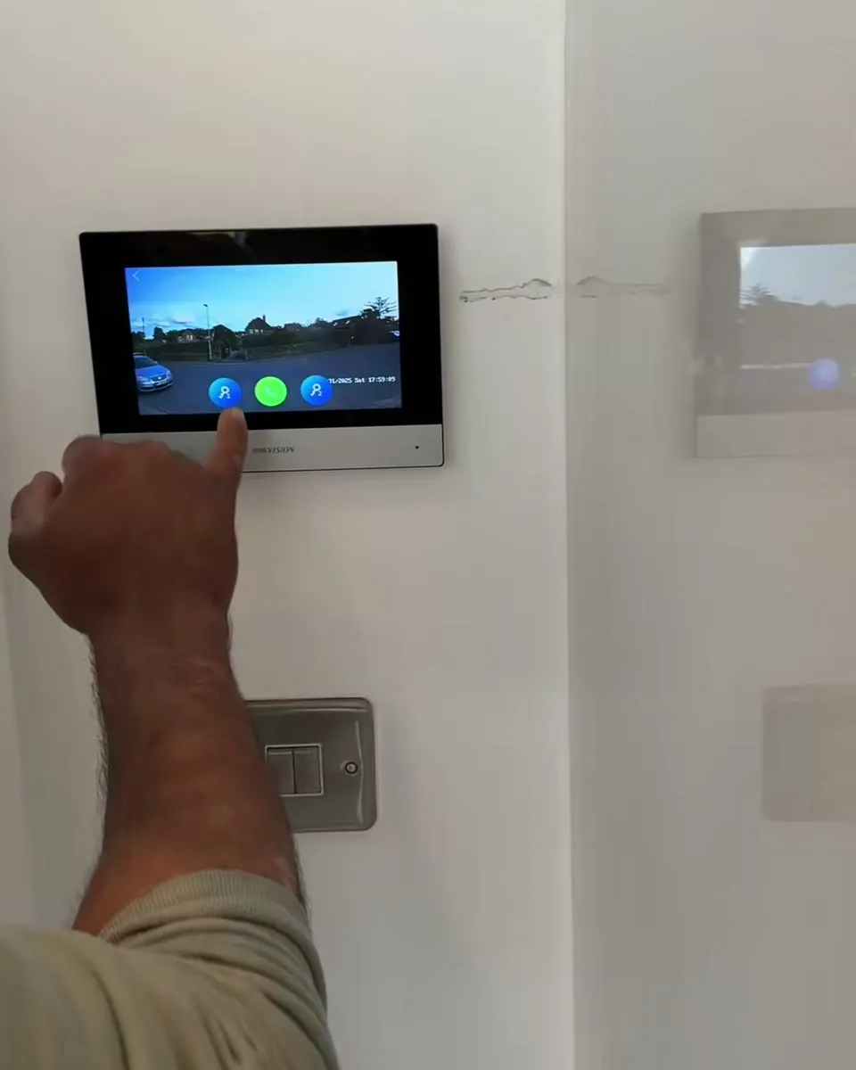 Mitu's hand pressing the screen of a Hikvision monitor on a white wall, showing the live view from a gate camera
