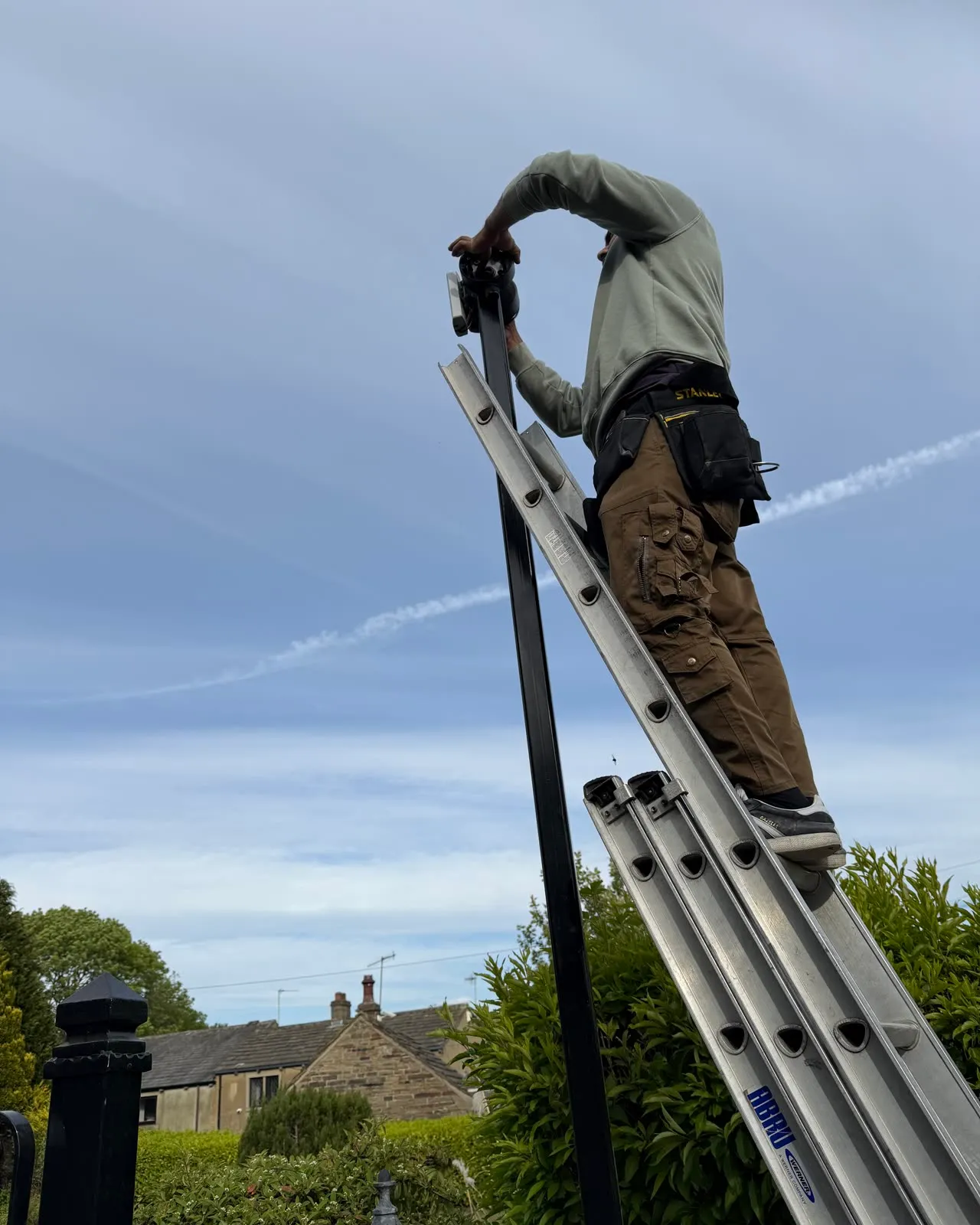 Mitu at the top of a long ladder against the sky, reaching up to fit a camera, stone houses behind him