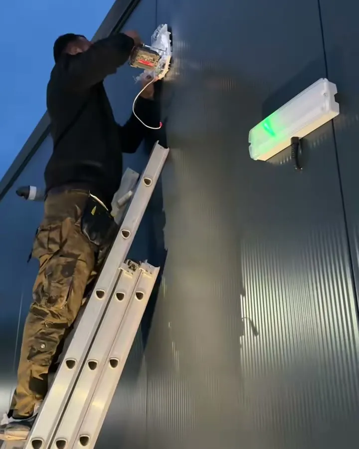Mitu up a ladder at dusk, drill in hand, fitting the siren base to the grey corrugated cladding of a shop