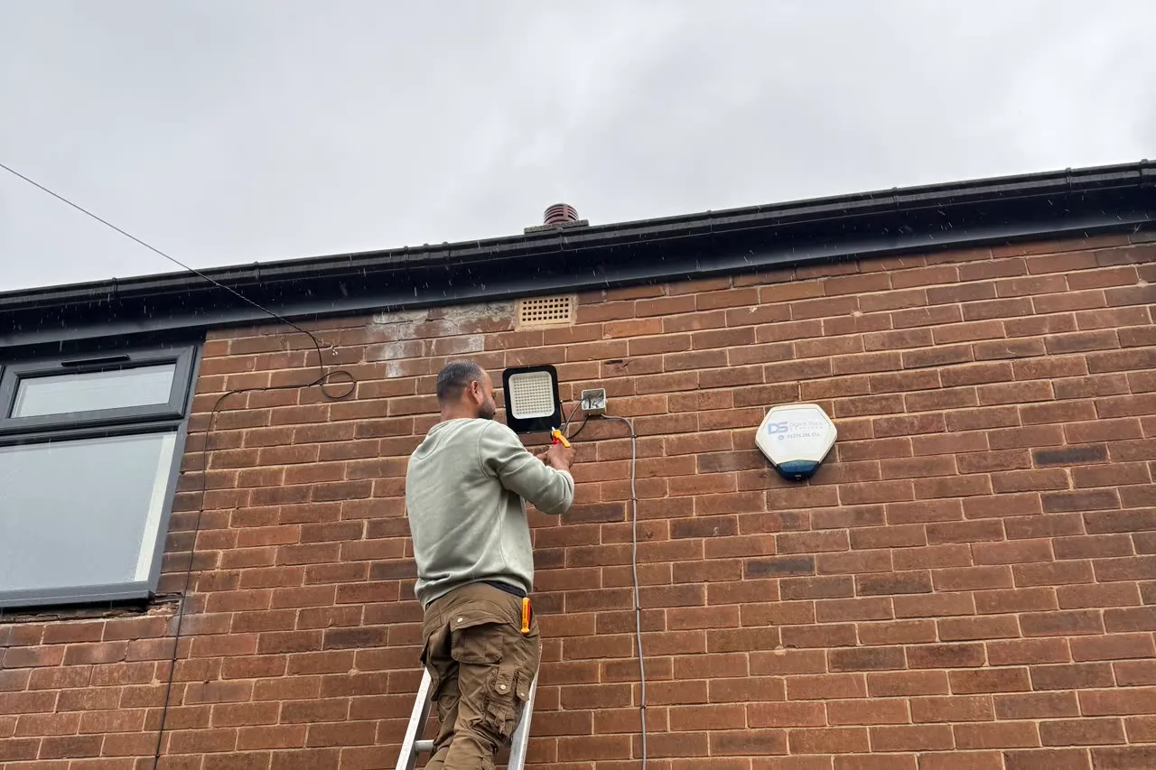 Mitu at the top of a ladder on a brick house, fixing a black LED floodlight to the wall beside the alarm bell box