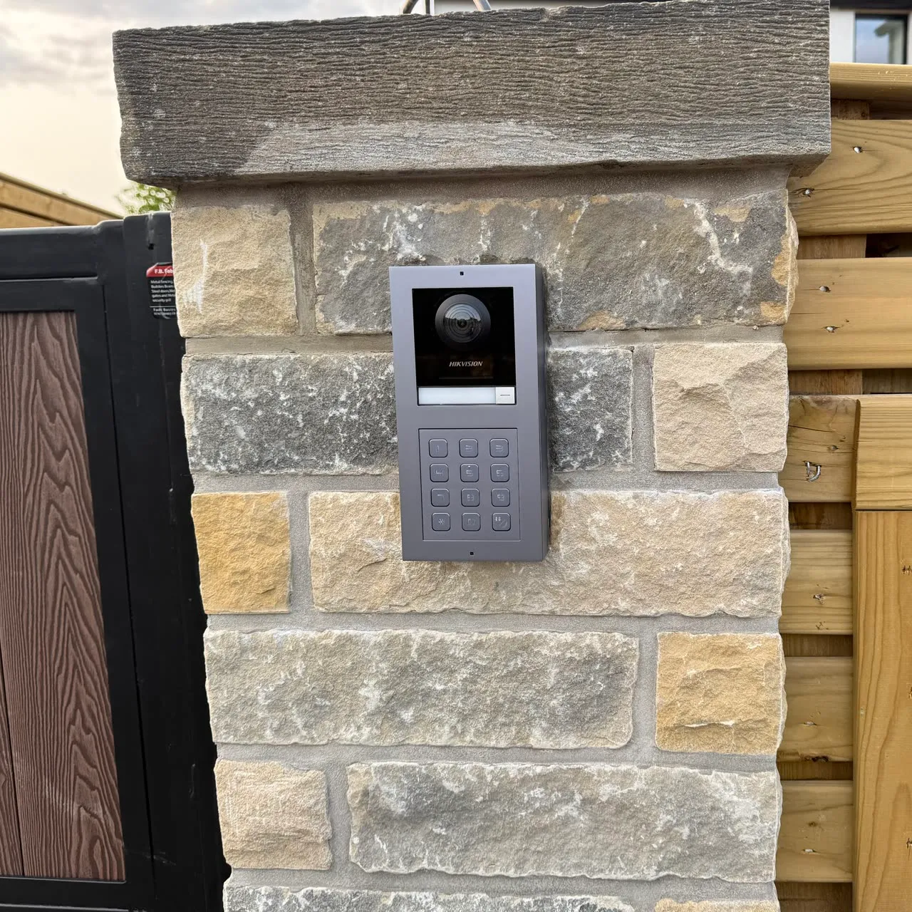 A Hikvision video intercom panel with keypad set into a stone gate pillar, the composite gate leaf beside it