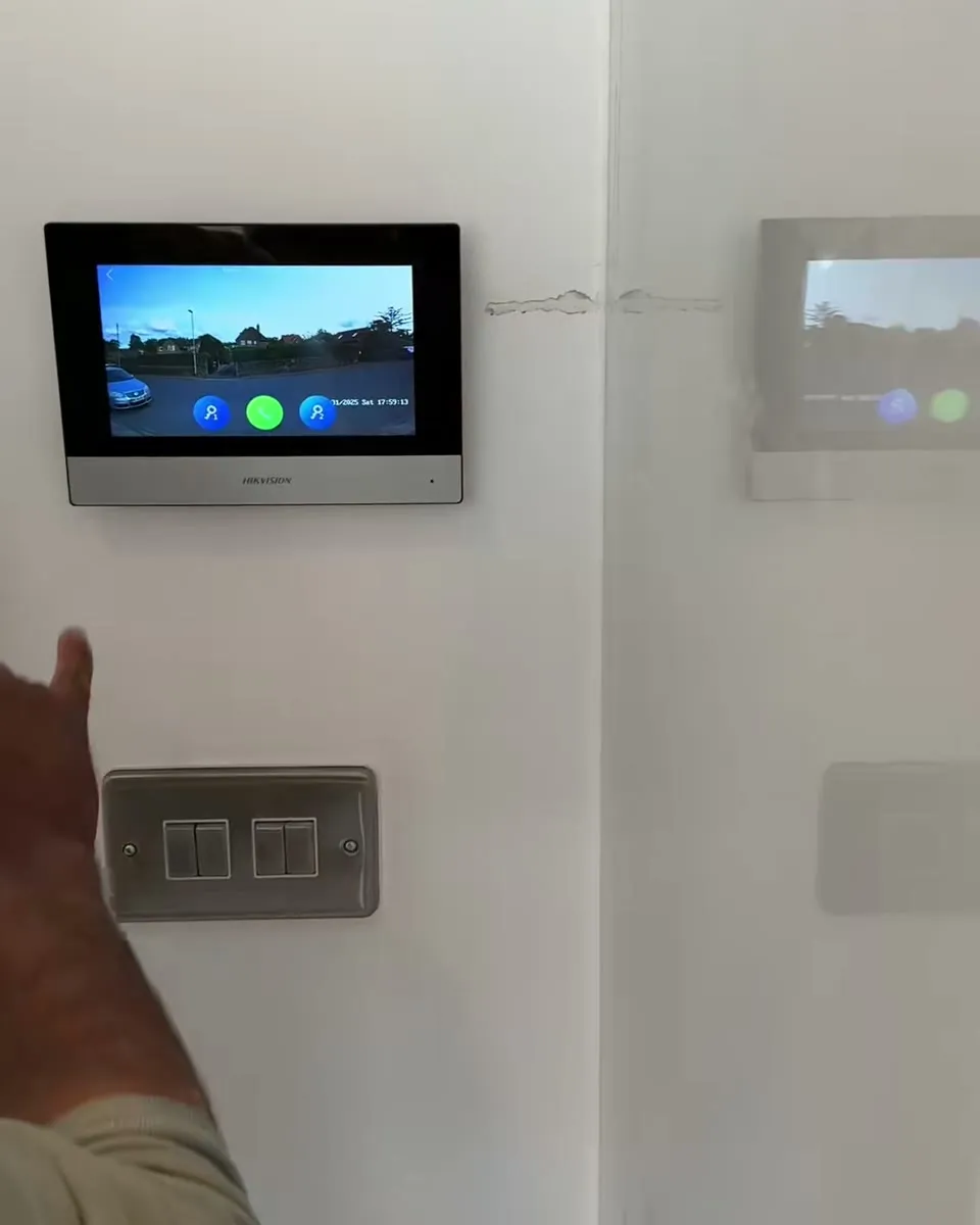 A Hikvision intercom monitor on a white wall inside the house, showing the driveway camera live