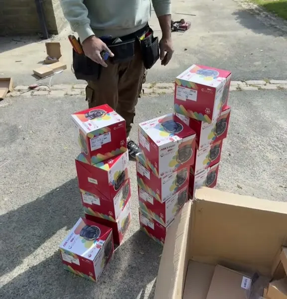 A stack of red Hikvision ColorVu camera boxes on a driveway, Mitu stood behind them with his tool belt on