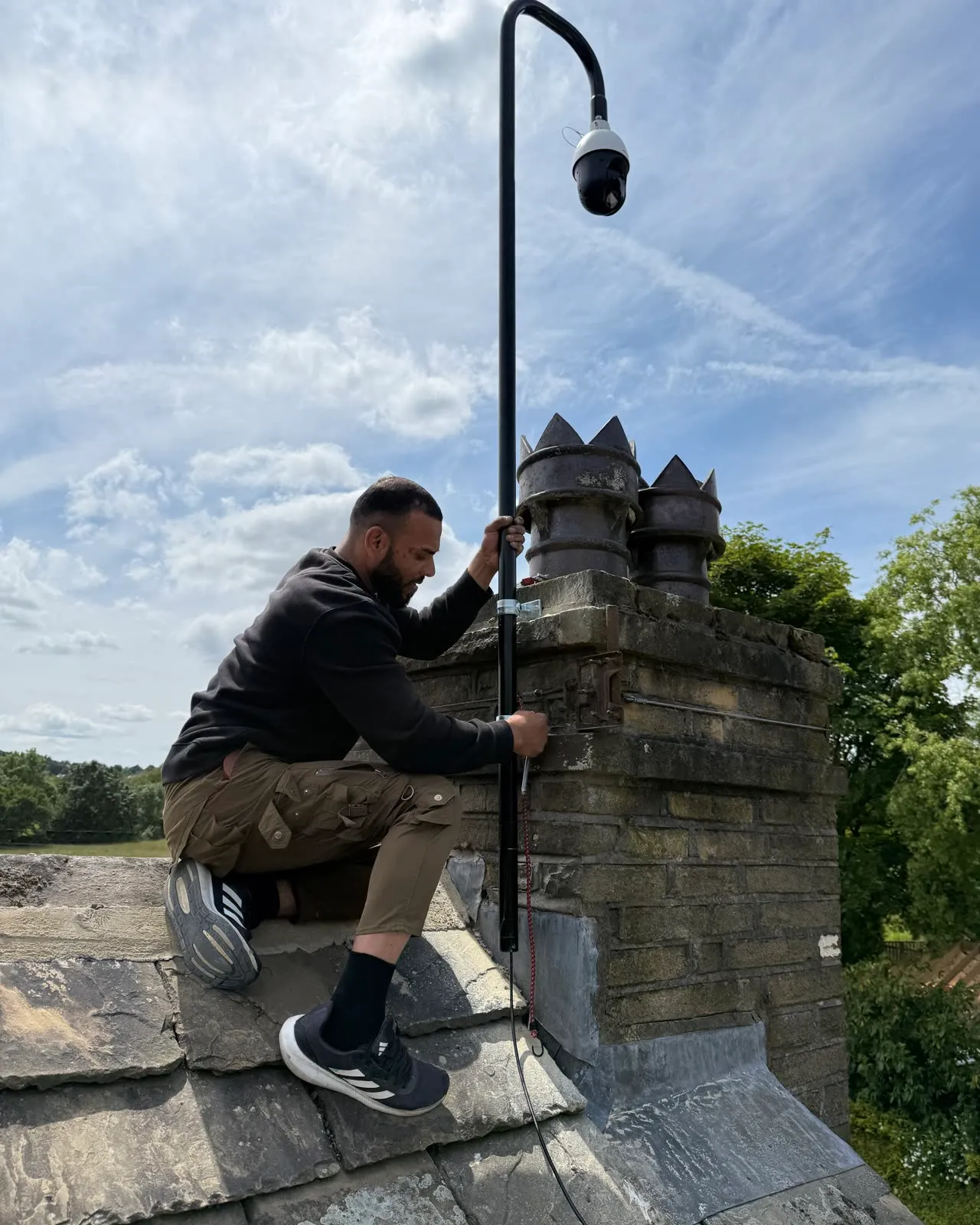 Mitu sat on the roof beside the chimney stack, fixing the curved pole bracket with the PTZ camera on top, against a bright sky