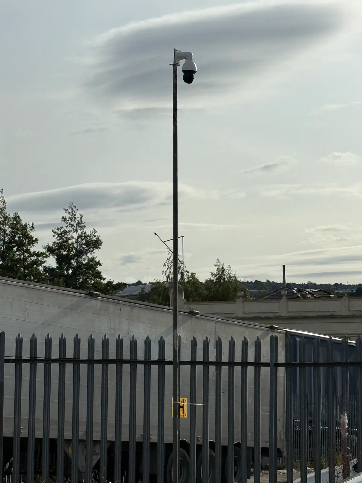 A Hikvision PTZ camera on a pole beside a stone chimney stack