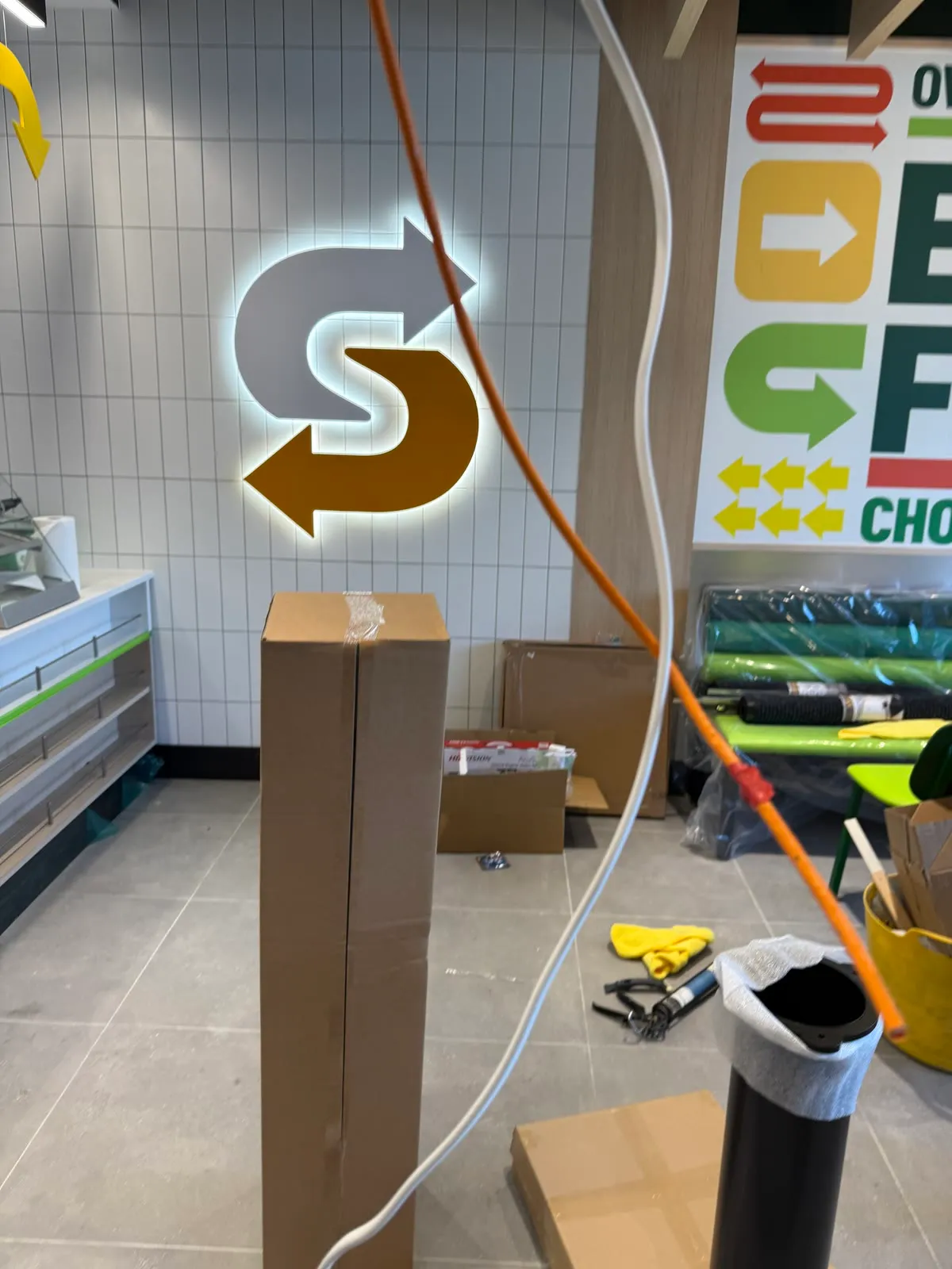 Inside the Subway mid-refit, the lit S logo on the tiled wall and a cable being run down beside it