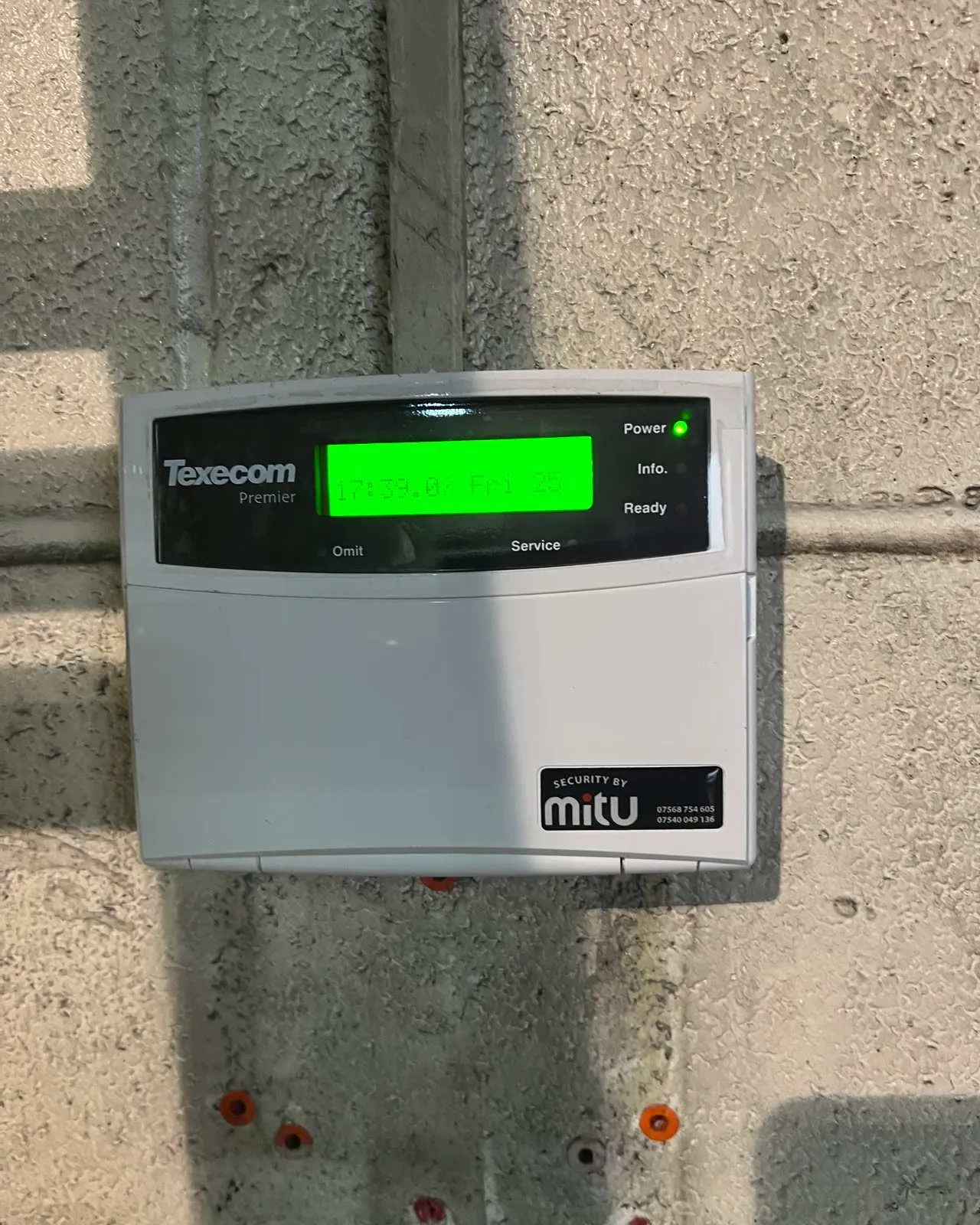 A Texecom Premier keypad on a bare wall with a Security By Mitu sticker in the corner, the display lit green