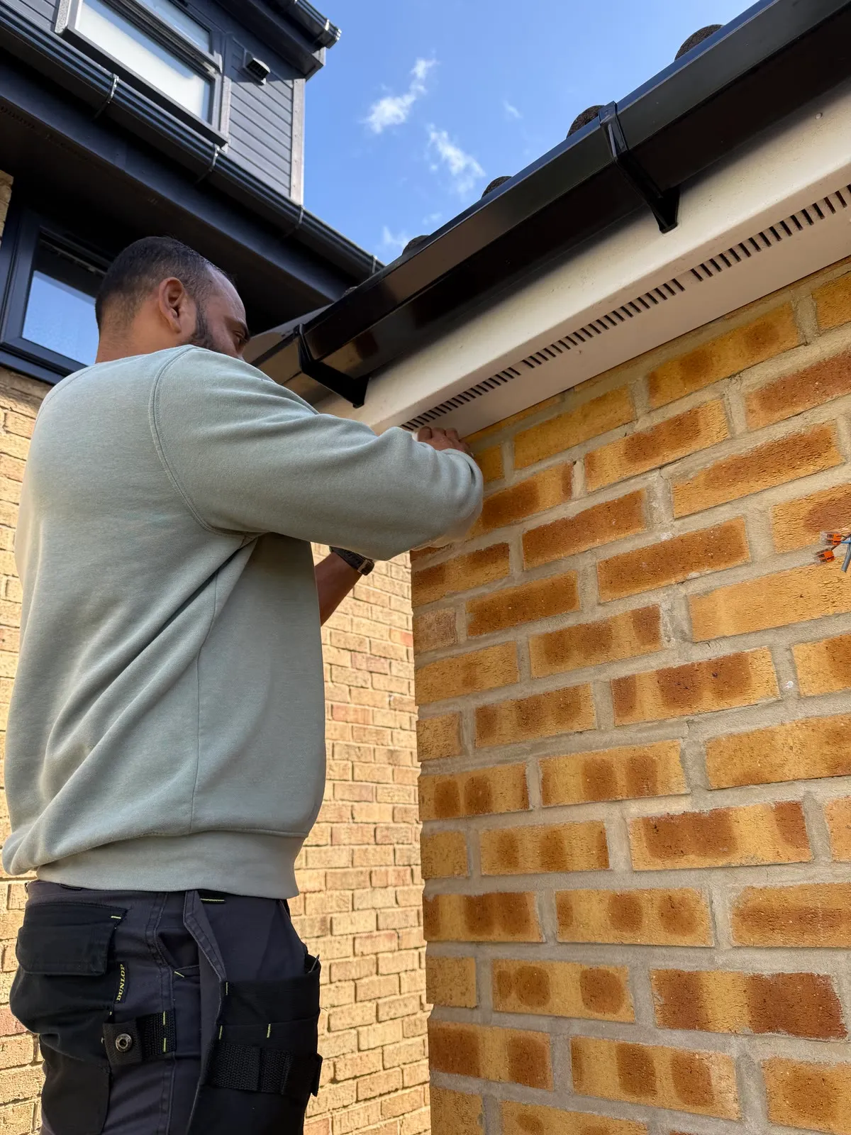 Mitu at the top of a ladder fitting a camera under the eaves of a red-brick house