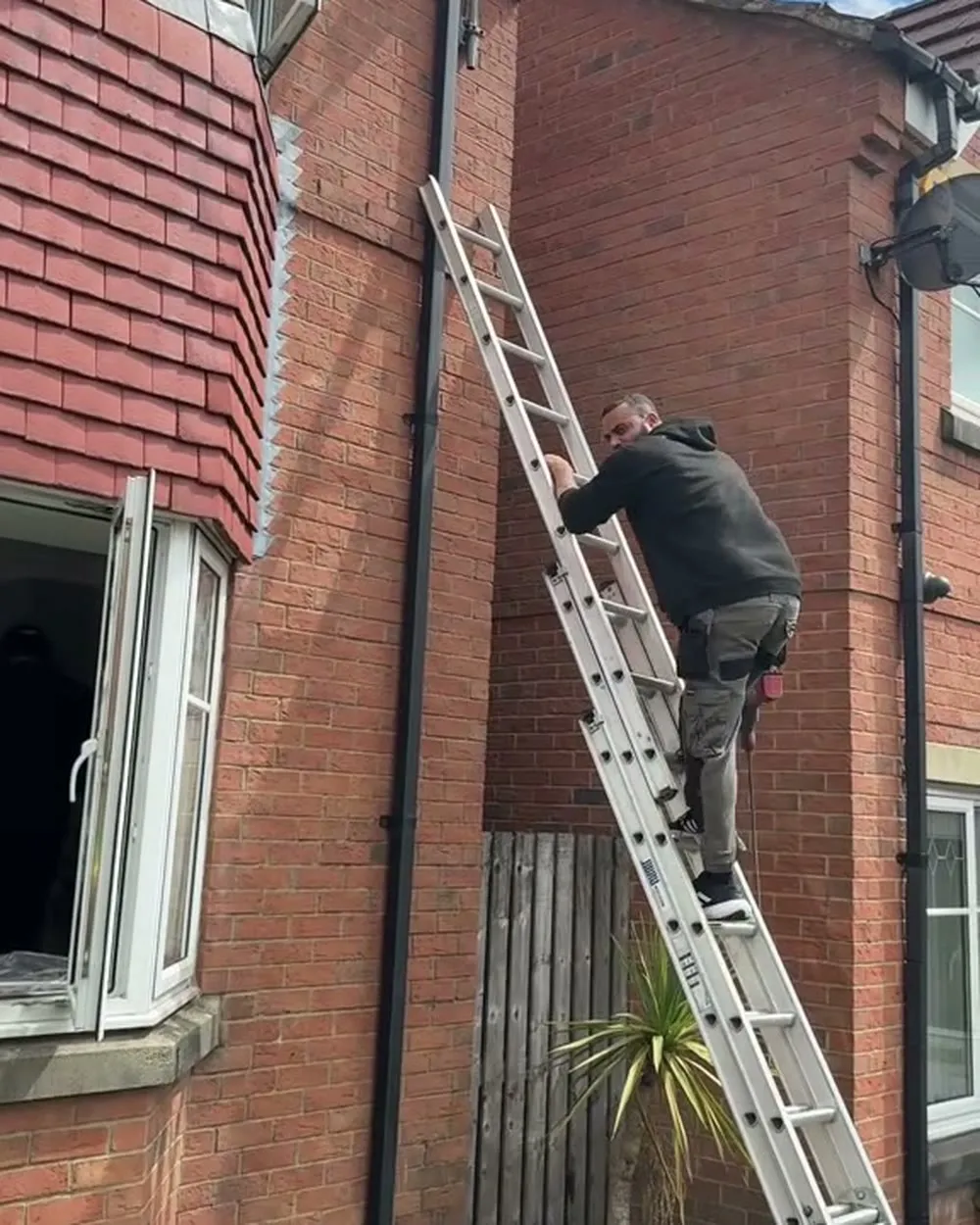 Mitu at the top of a ladder fitting a camera under the eaves of a red-brick house