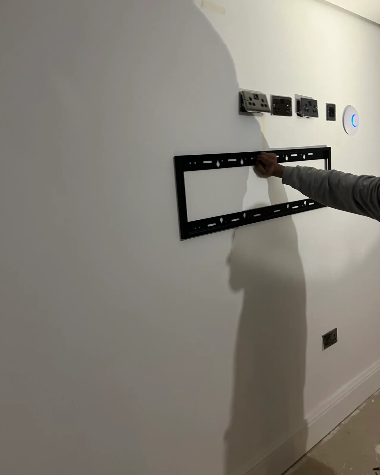 Mitu holding a TV wall bracket level against a freshly plastered wall, the double socket and data point already fitted where the screen will cover them