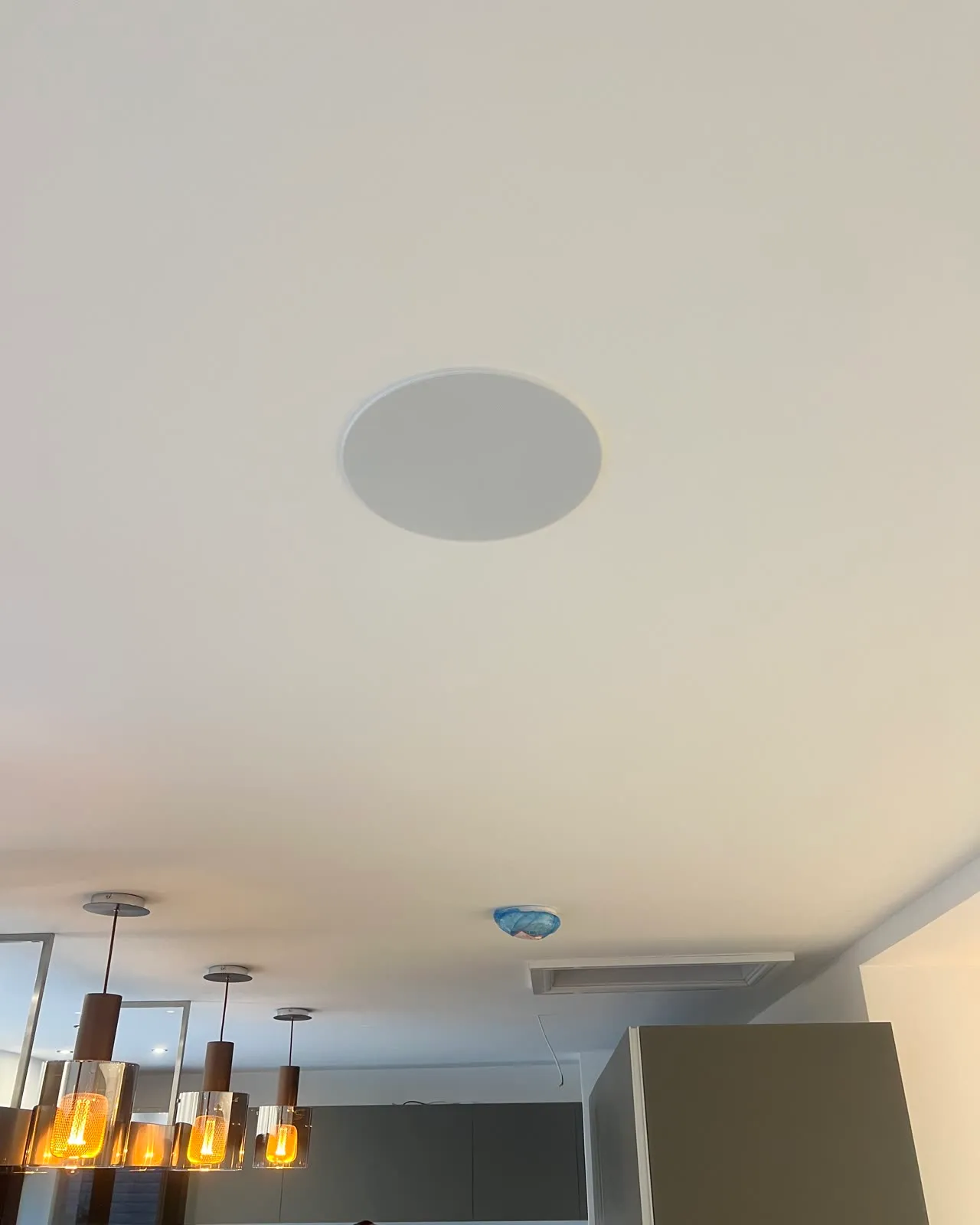 A round in-ceiling speaker set flush into a white kitchen ceiling, pendant lights below it