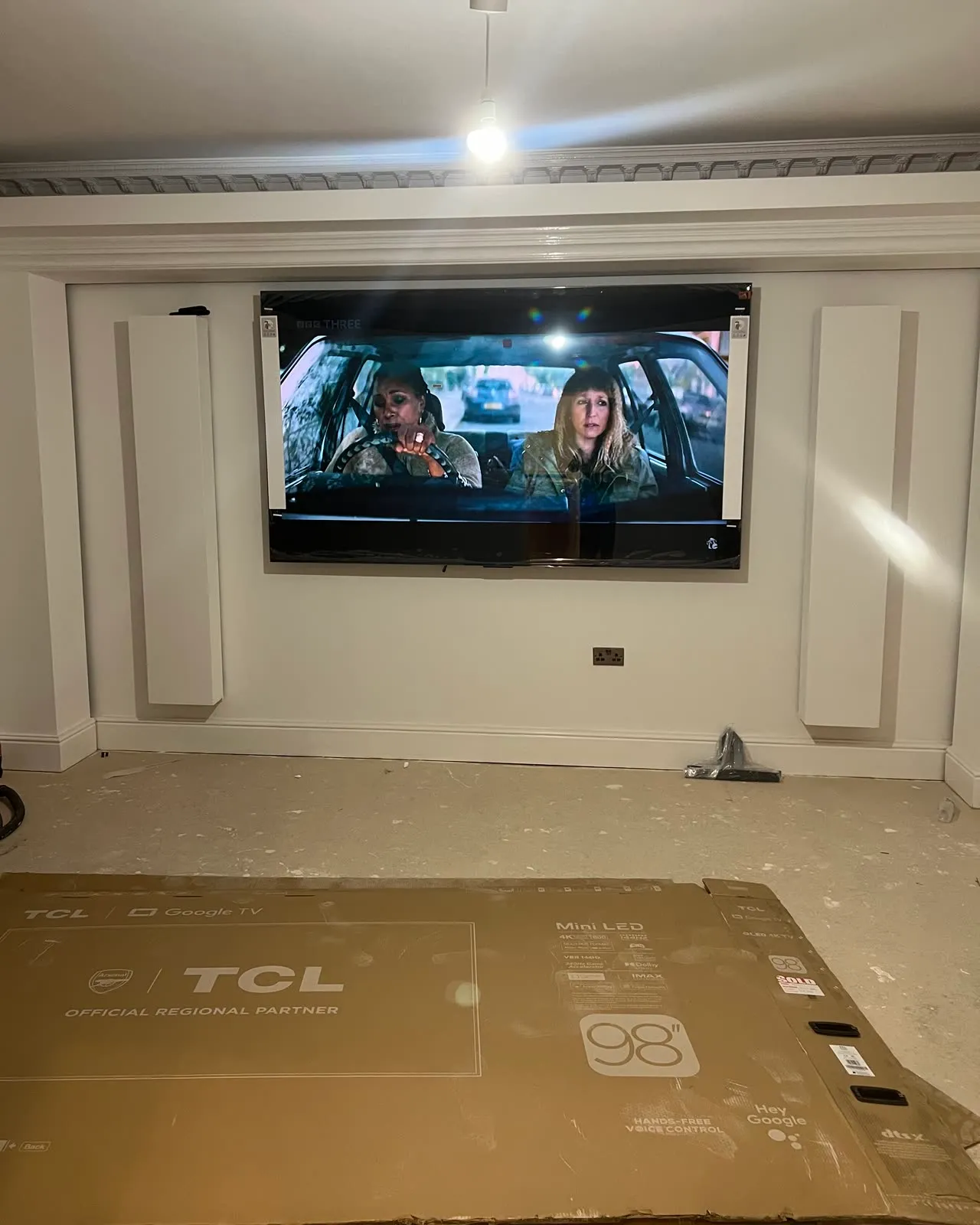 A finished cinema room: a large TV mounted flush on a panelled wall between two columns, no cable in sight, the floor still covered while the room is finished