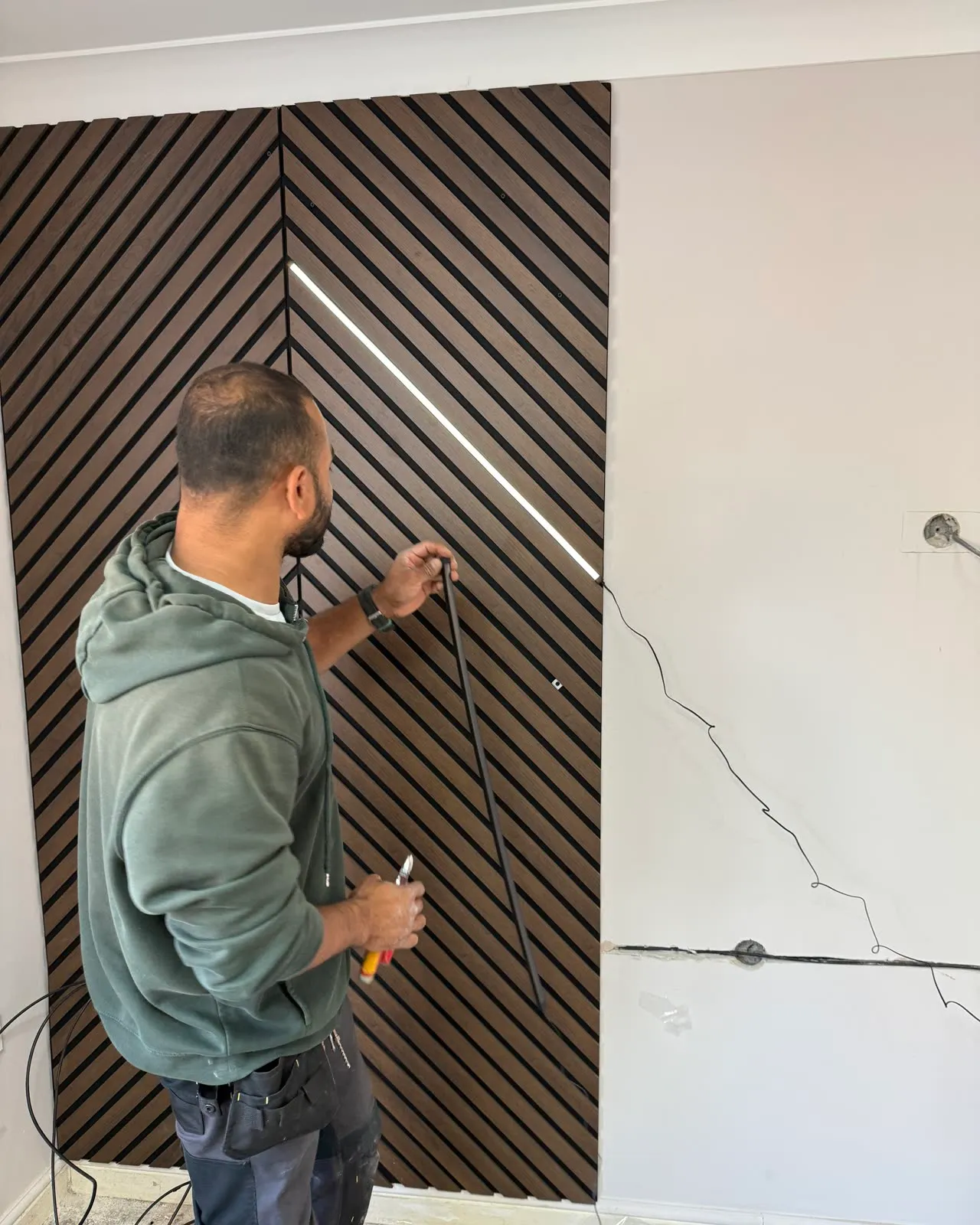 Mitu fitting a slim LED light profile into a dark slatted wall panel, the wiring for it coming out of the wall beside the panel